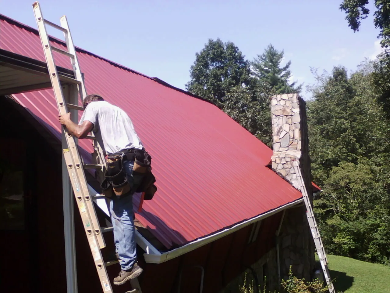 Roofer on a ladder beside a red metal roof and stone chimney — Soffit & Fascia Repair by Roofing Brothers in Somersworth